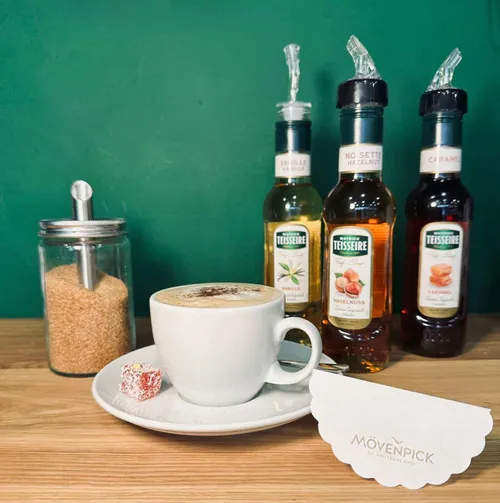 Ein Capuccino mit verschiedenen Syrupsorten im Hintergrund